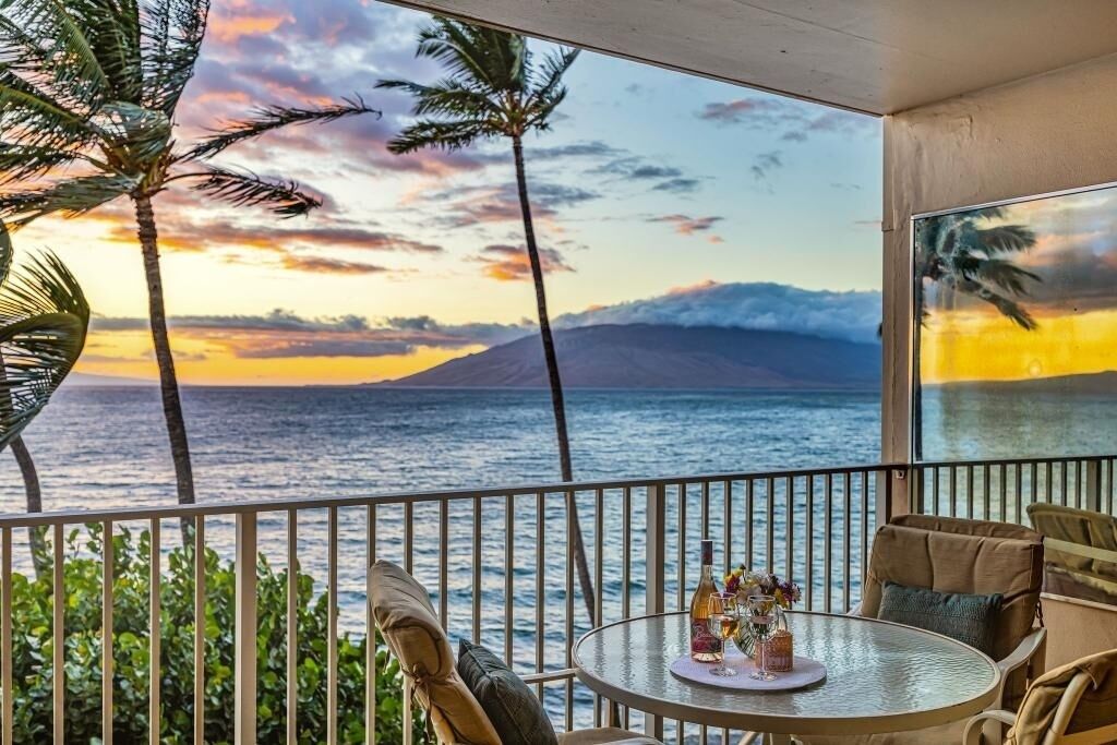 A balcony with a view of the ocean and a sunset. There is a round table with a bottle and flowers on it.