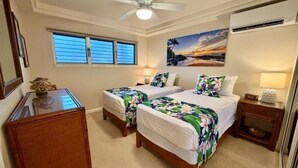 A hotel room with two beds, a ceiling fan, a window with blinds, a painting of a beach scene, and a small table with a lamp. - Royal Mauian, #515 2 Bedroom Condo (Kihei)