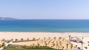 Una playa con vista despejada al mar, una zona arenosa con árboles y algunos edificios a lo lejos.