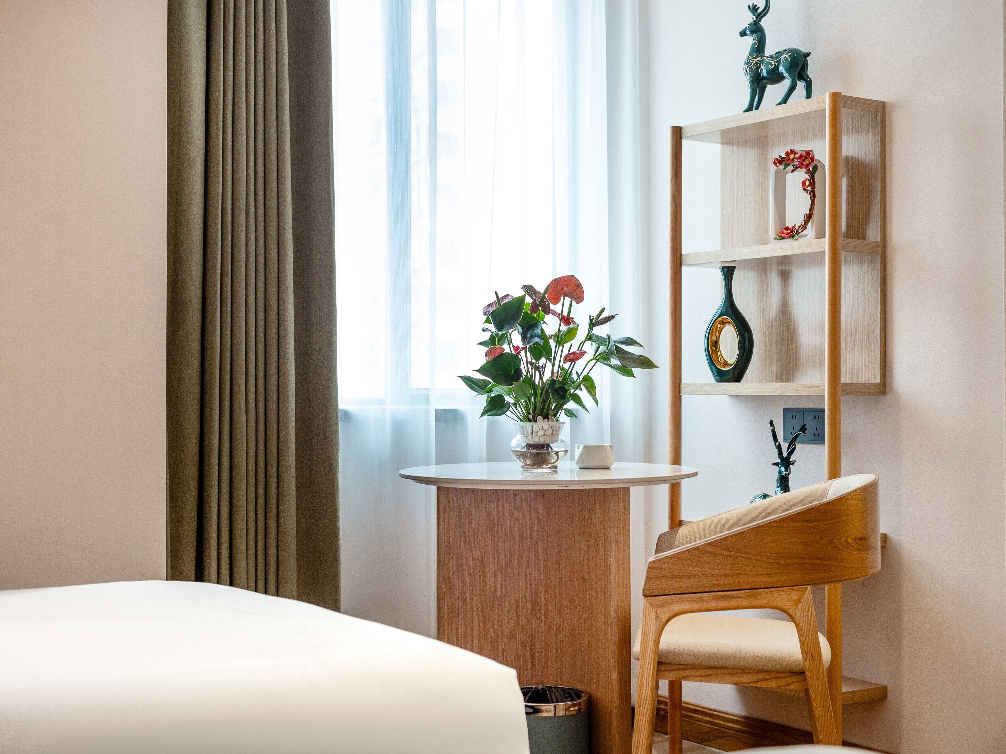 Una habitación de hotel con una cama, una silla de madera, una mesita con una planta y una estantería con adornos.
