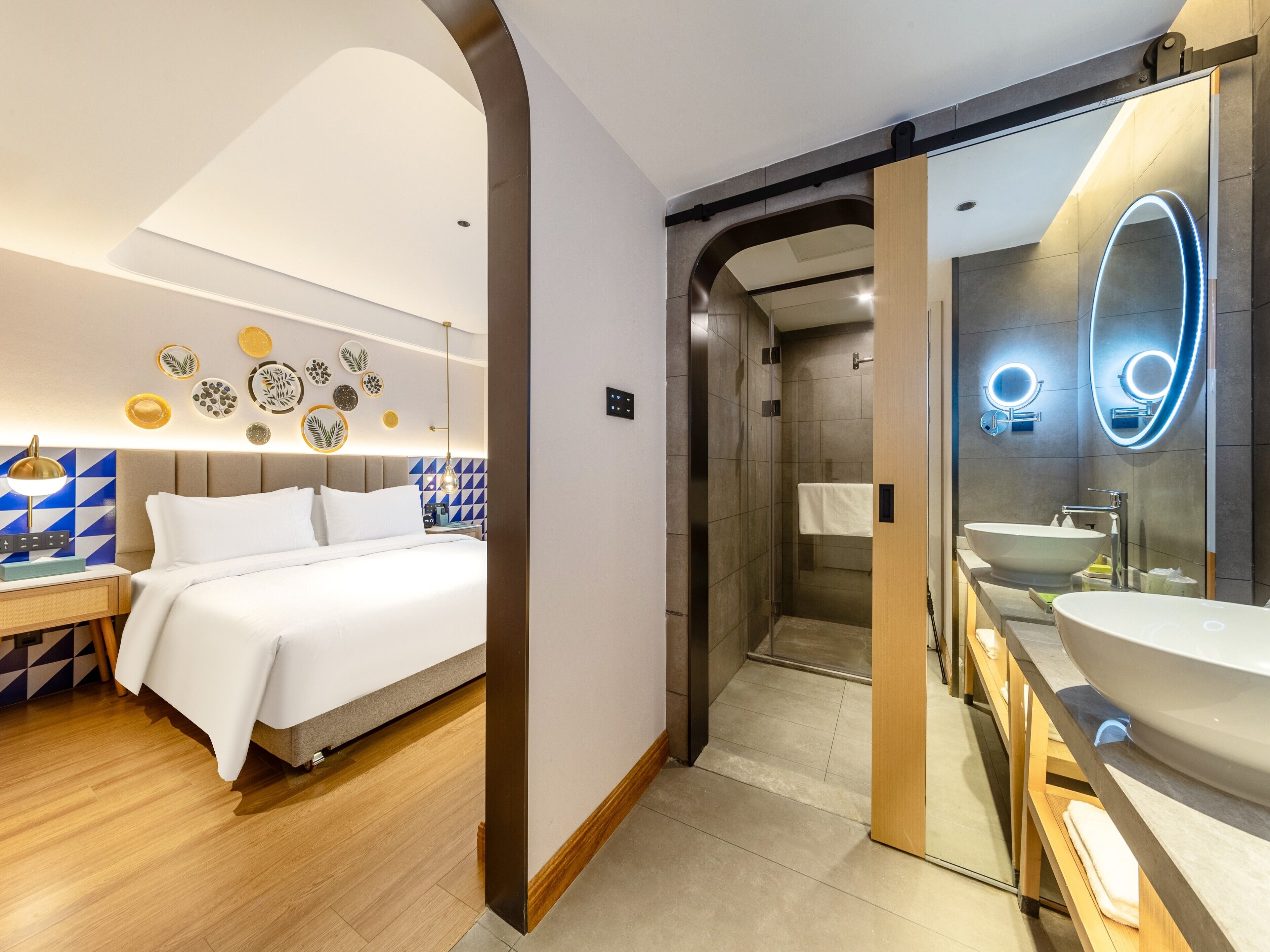 A modern hotel room with a bed, bedside table, and a bathroom with a large mirror and sink.