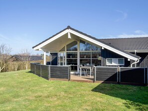Una casa moderna con un amplio ventanal, un patio con mobiliario exterior y un techo inclinado.