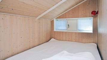 Una habitación con paredes de madera, techo inclinado, cama con ropa blanca y ventana con persianas.