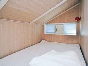 A room with wooden walls, a sloped ceiling, a bed with white bedding, and a window with blinds.