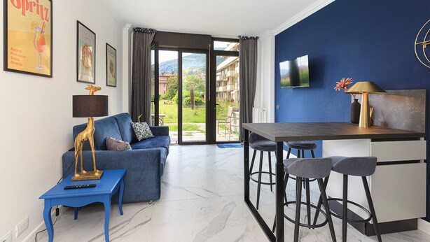 A modern living room with a blue sofa, a small table, and a kitchen area with bar stools. - Dolce Vita Baveno (Baveno)