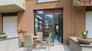 A balcony with wicker furniture and potted plants, a sliding glass door leading to a covered outdoor area, and a mountain view in the background. - Dolce Vita Baveno (Baveno)