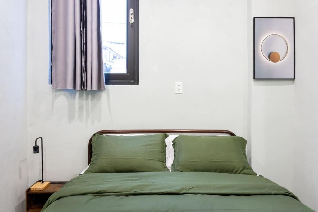 Une chambre à coucher comprenant un lit, une table de chevet, une lampe fixée au mur, une fenêtre avec des rideaux et un tableau encadré.