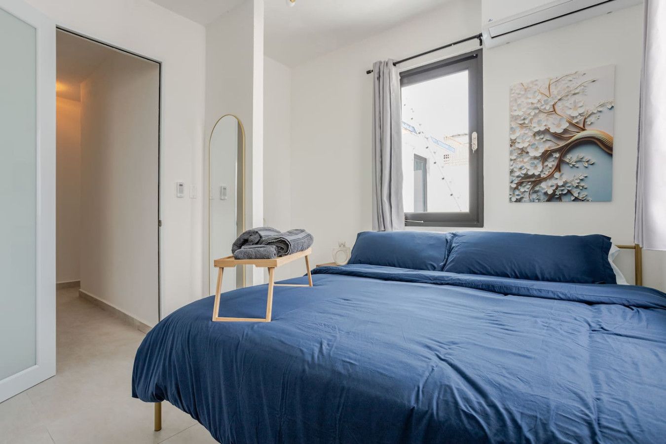 A bedroom with a large bed, a wooden bedside table with towels, a window with curtains, and a wall-mounted artwork.
