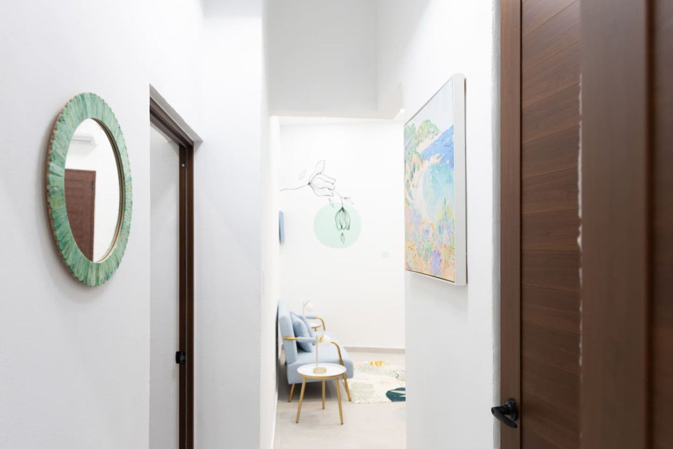 A hallway with a mirror, a door, and a room with a chair and a table.