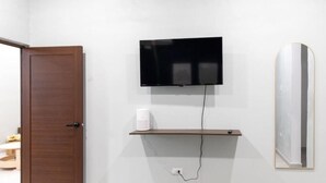 A room with a wooden door, a mounted television, a white coffee maker on a shelf, and a mirror with a gold frame.