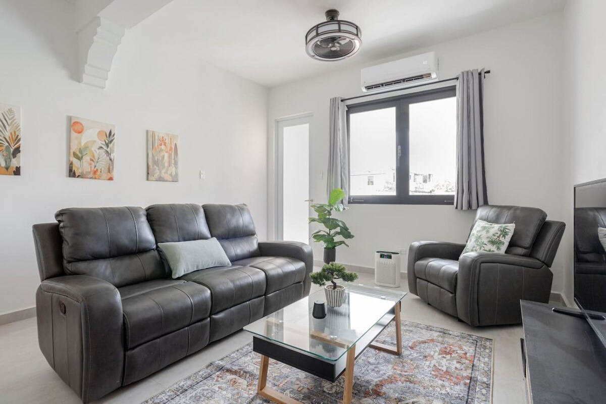 Een moderne woonkamer met een leren bank, een glazen salontafel en een grijze fauteuil. Er is een groot raam met gordijnen, een plafondventilator en een airconditioningsysteem.