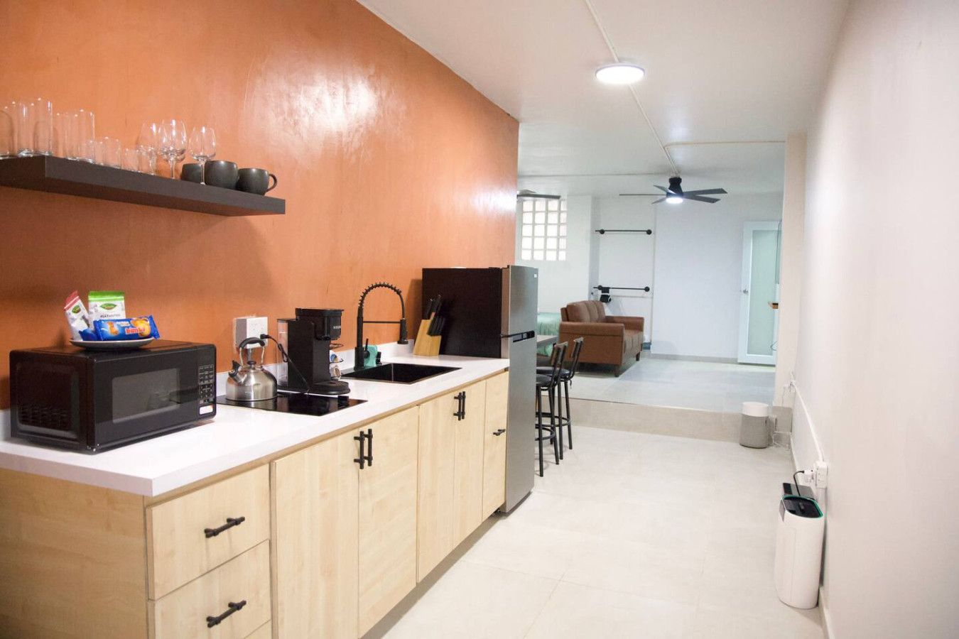 A compact kitchen with a microwave, coffee maker, and a sink. There is a shelf with glasses and a bag of snacks. A ceiling fan is visible in the background.