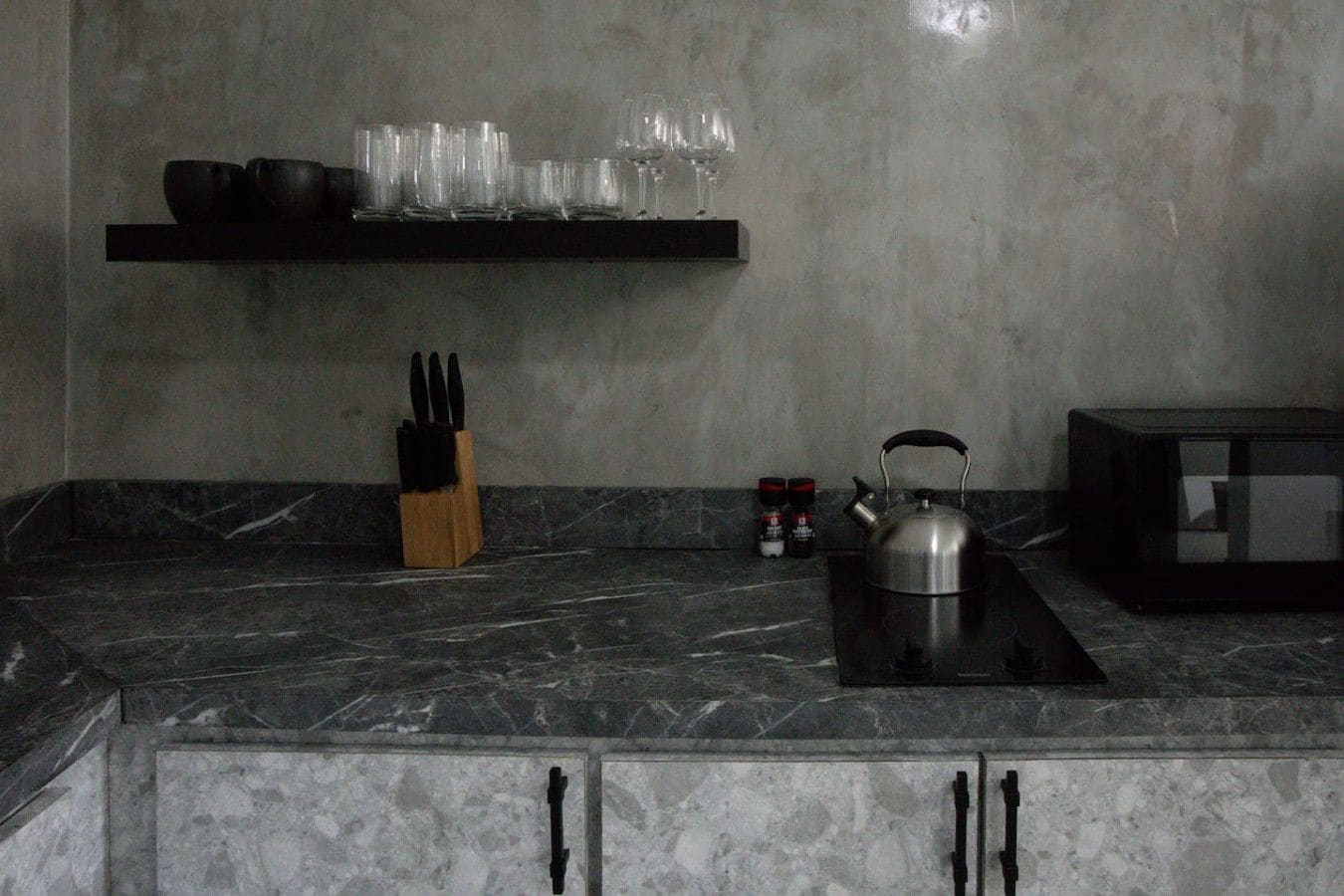 Una cocina con encimera de mármol, hervidor, bloque de cuchillos y estante con vasos y cuencos.