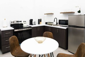 A modern kitchen with dark cabinetry, a stainless steel refrigerator, a microwave, and a kettle on the counter. There is a white table with a wire basket in the center and two brown chairs. - Casa Cielo (San Juan)