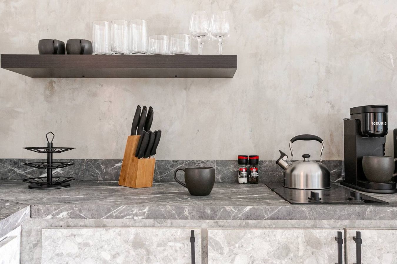 A kitchen with a marble countertop, a coffee maker, a kettle, a knife block, a spice rack, and a shelf with glasses and cups.