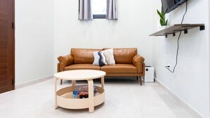 A living room with a brown leather sofa, a round wooden coffee table, a flat-screen TV mounted on the wall, and a small potted plant on a shelf.