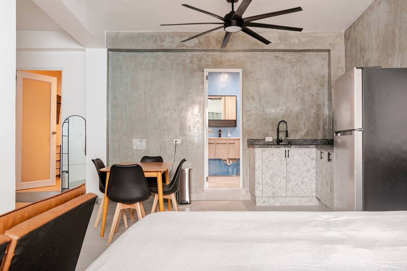 A modern hotel room with a bed, a dining table with chairs, a kitchenette, and a ceiling fan.