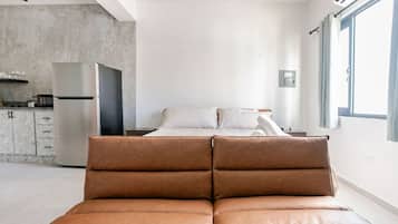 Un dormitorio moderno con cama, un sofá en primer plano, una zona de cocina y un ventanal grande.