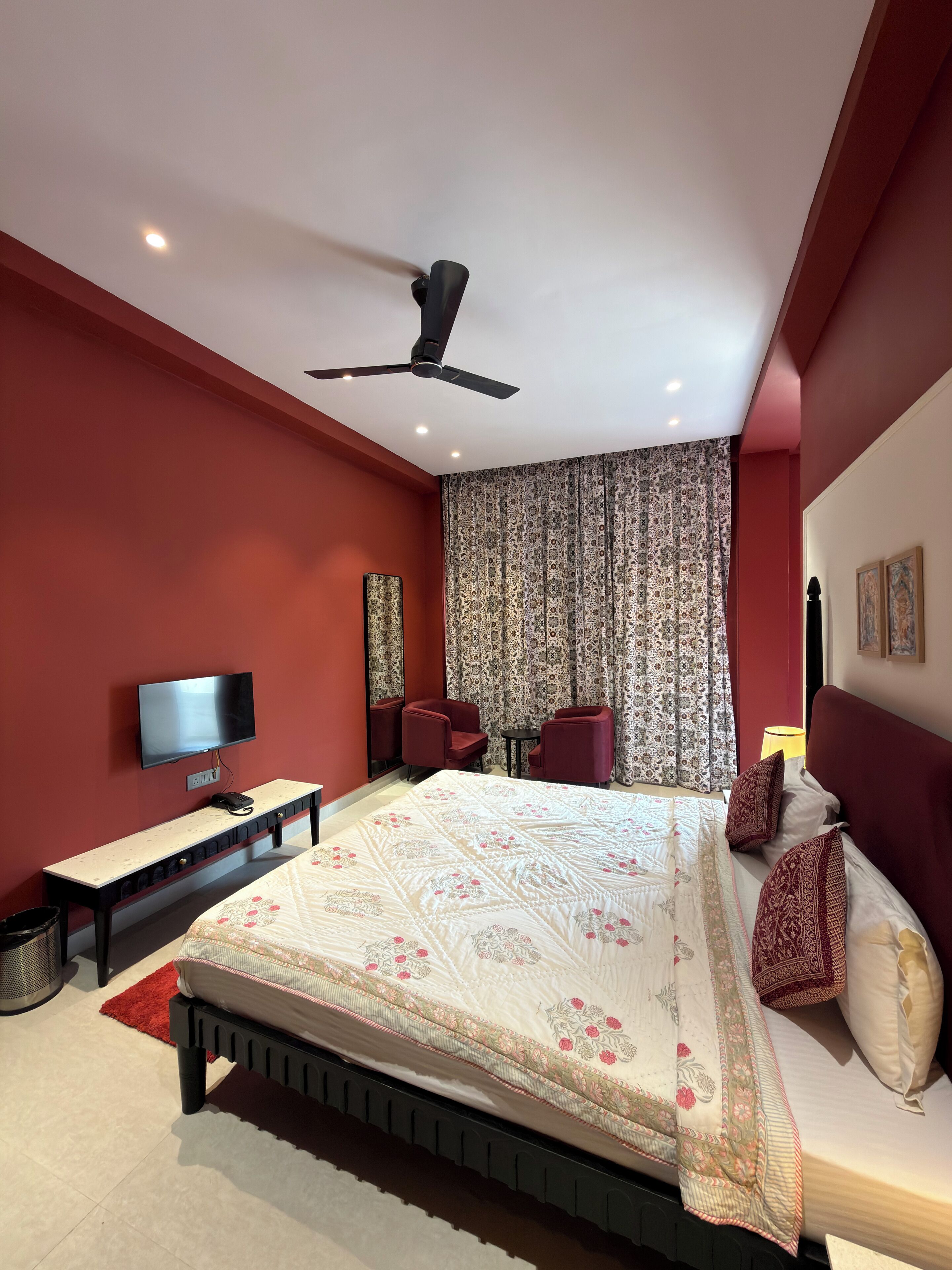 A bedroom with a bed, a ceiling fan, a TV, two chairs, a desk, and a patterned curtain.