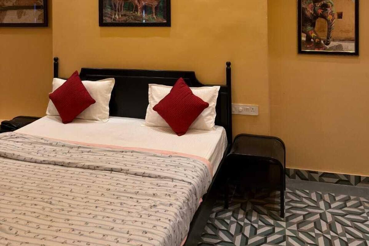 A hotel room with a bed, two red pillows, a nightstand, and a wall-mounted painting of deer.