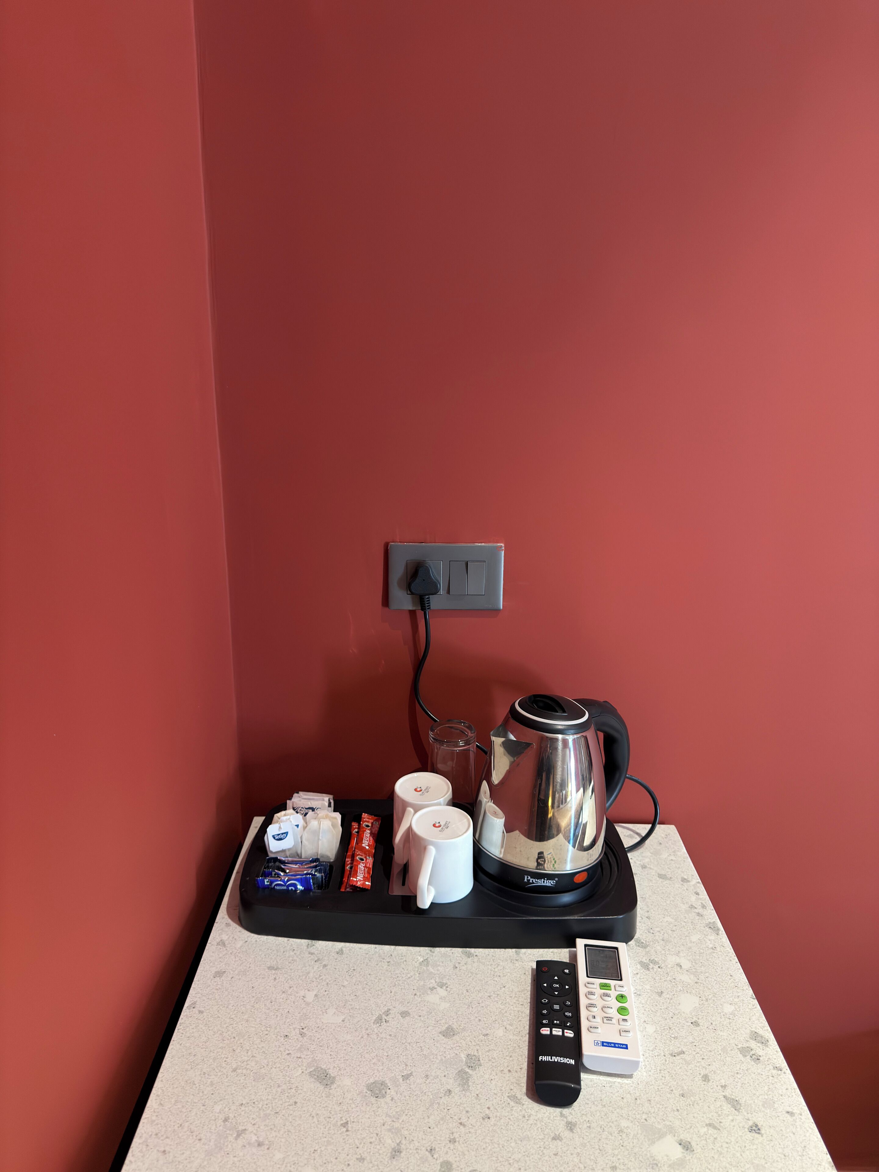 A kitchenette with a coffee maker, a kettle, a remote control, and some snacks on a countertop.