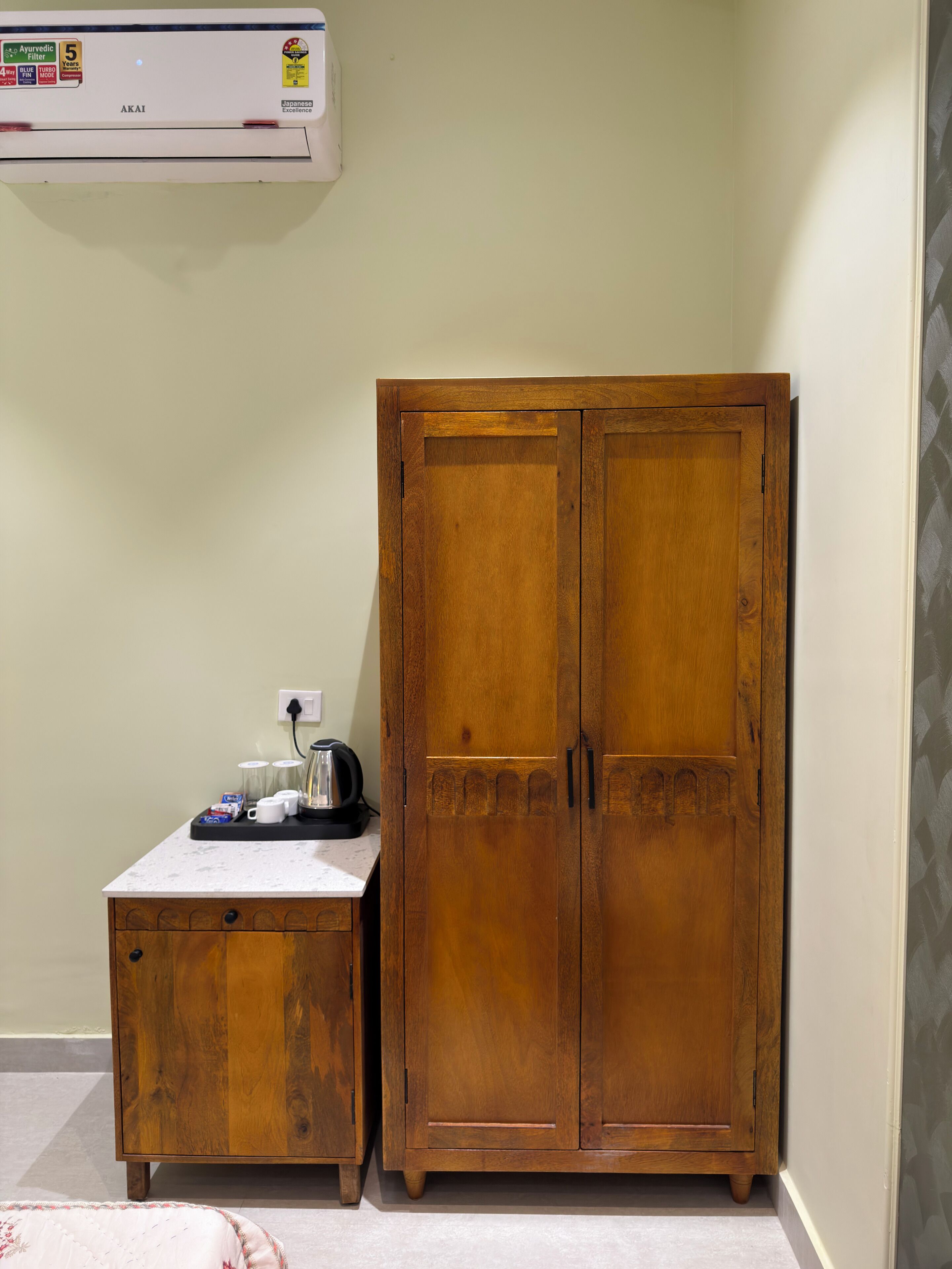 A room with a wooden wardrobe, a small table with a kettle and cups, and an air conditioner on the wall.