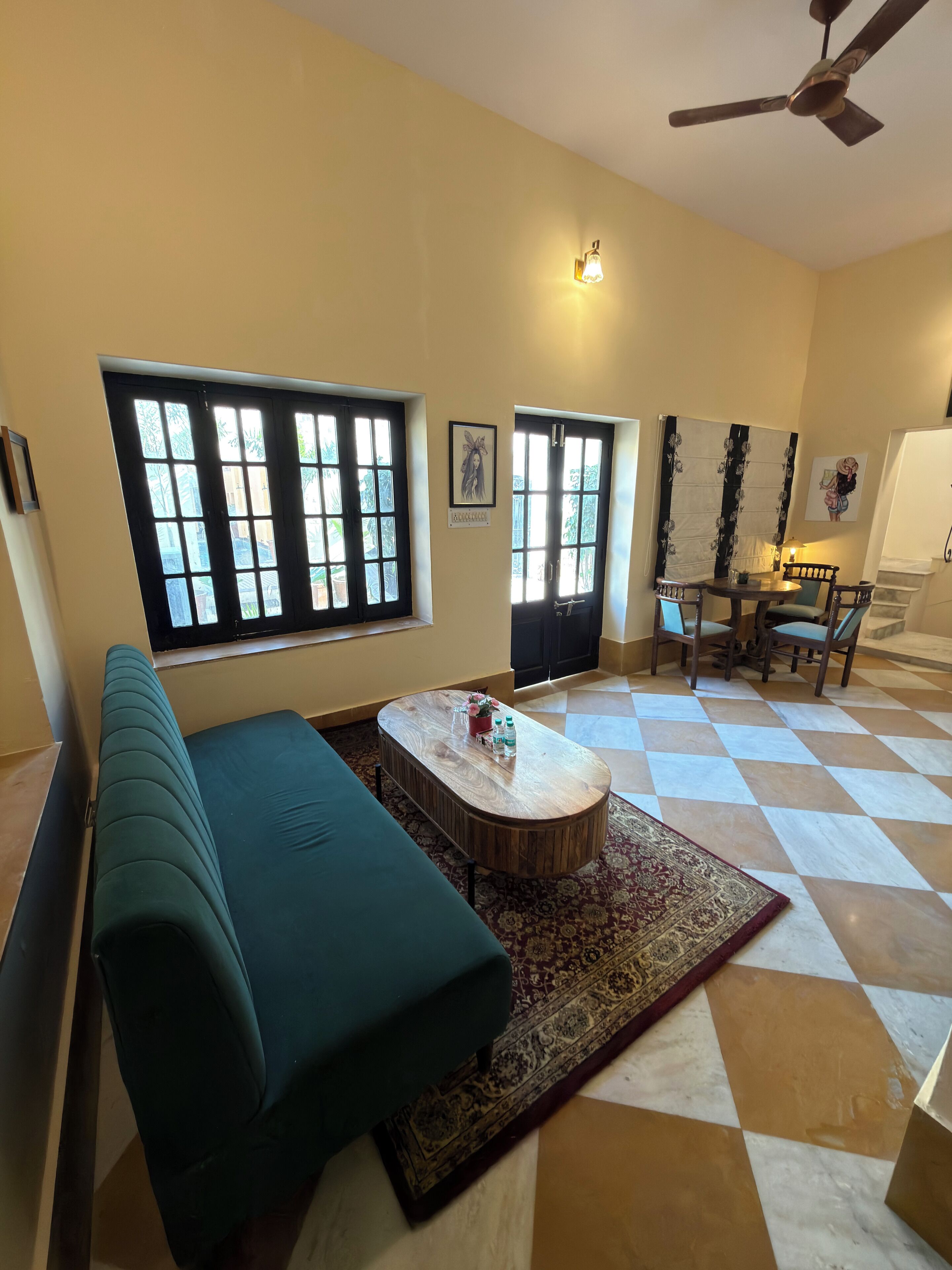 A living room with a green sofa, a wooden coffee table, and a patterned rug. There are two windows with black frames and a ceiling fan.