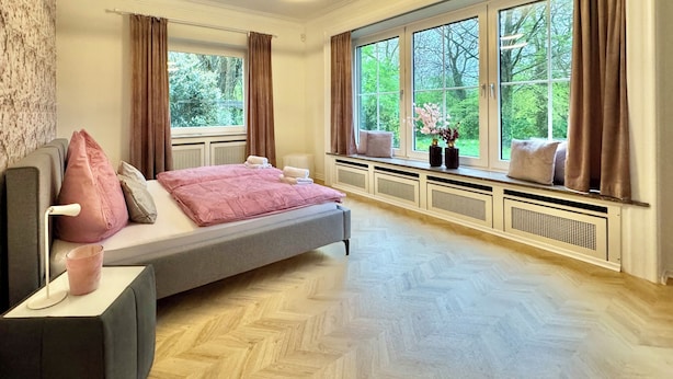 A bedroom with a bed, a bedside table, a window with curtains, and a view of greenery outside.