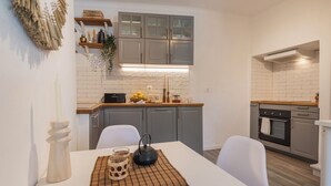 Μια μοντέρνα κουζίνα με γκρι ντουλάπια, έναν λευκό πάγκο και έναν εντοιχισμένο φούρνο. Υπάρχει ένα τραπέζι φαγητού με ένα σουπλά και μια βάση για κερί.
