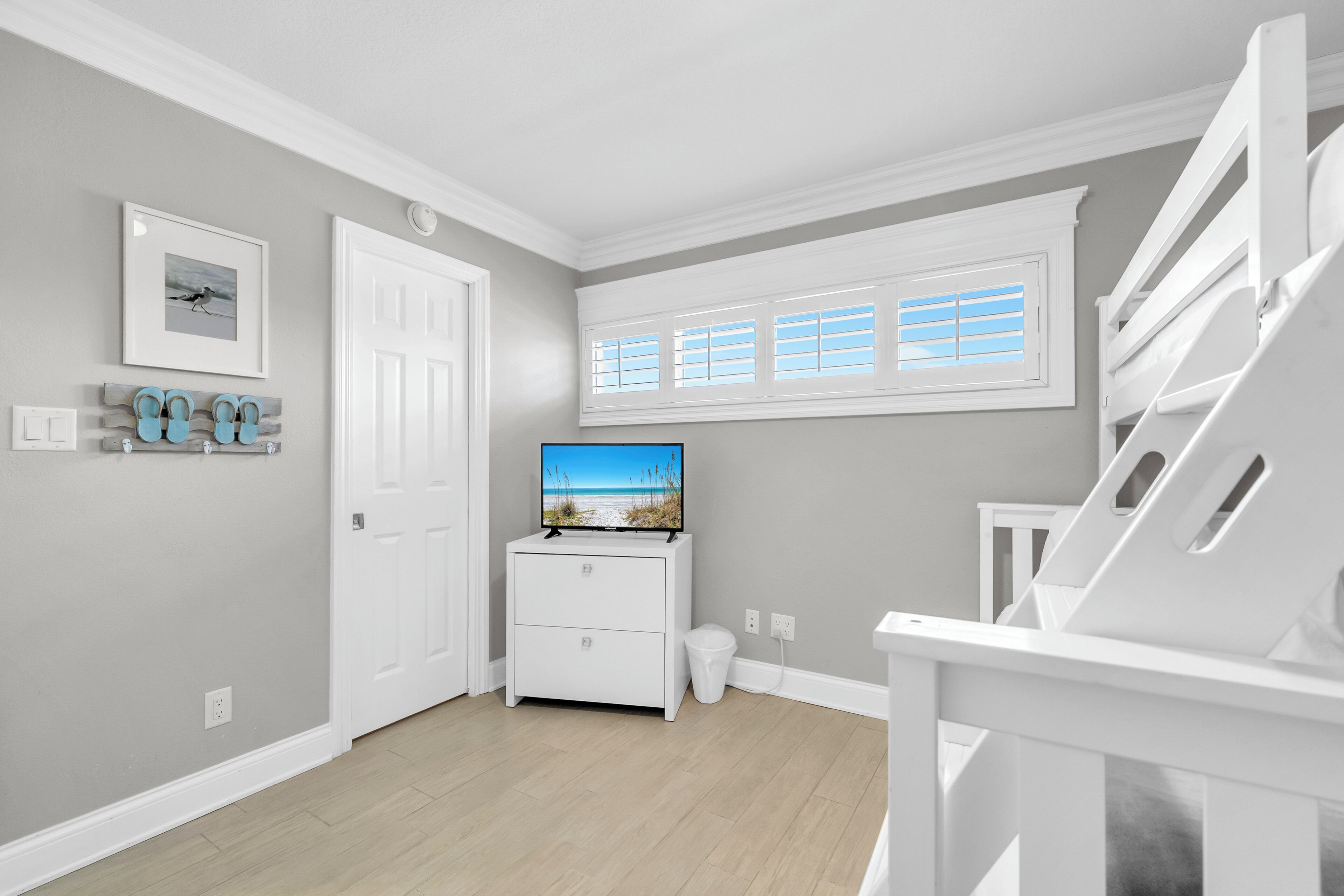 A room with a white door, a white dresser, a television on top, a window with blinds, and a staircase.