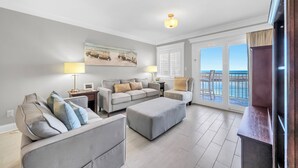 A modern living room with a grey sofa set, a coffee table, and a TV stand. There is a large window with a view of the sea.