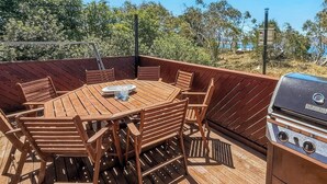 Una terraza al aire libre con una mesa y sillas de madera, una parrilla y vistas a árboles y un cuerpo de agua.