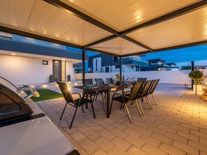 Un patio exterior con zona de comedor cubierta, un coche y un edificio moderno al fondo.