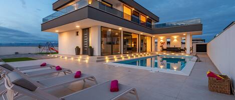A modern two-story house with a swimming pool, outdoor seating, and a view of the mountains.