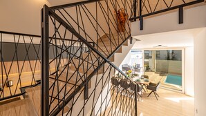 Un intérieur moderne avec un escalier doté d’une rampe en métal noir, un coin repas avec une table et des chaises, et une vue sur une piscine à travers des portes vitrées.