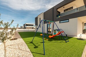 Ein Spielplatz mit Schaukeln und Rutsche, umgeben von einem Gebäude mit großen Fenstern und einem Kiesweg.