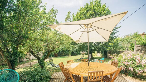 Ein runder Holztisch mit Stühlen und einem großen, beige gefärbten Sonnenschirm in einem Garten.