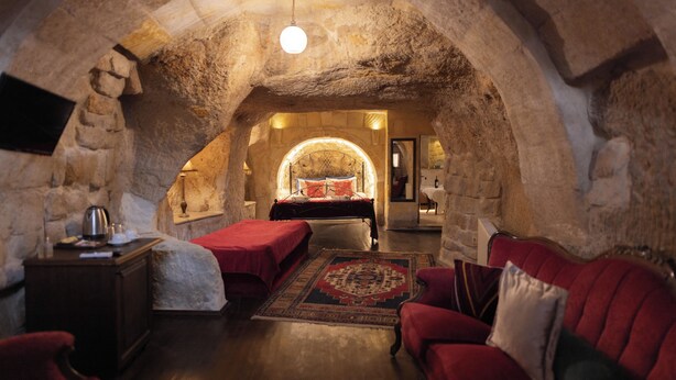 A cozy, stone-walled room with a bed, a red sofa, and a small table. - roca cappadocia suites (Ürgüp)