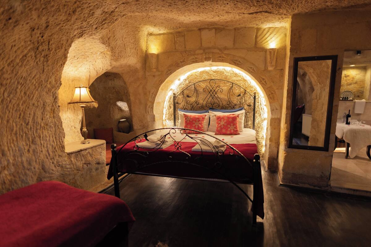 A bedroom with a stone wall, a bed with red bedding, a nightstand with a lamp, and a mirror on the wall.