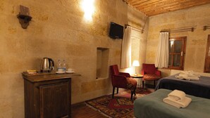 A room with a wooden cabinet, a red chair, a bed with a blue bedspread, and a small table with a lamp. - roca cappadocia suites (Ürgüp)