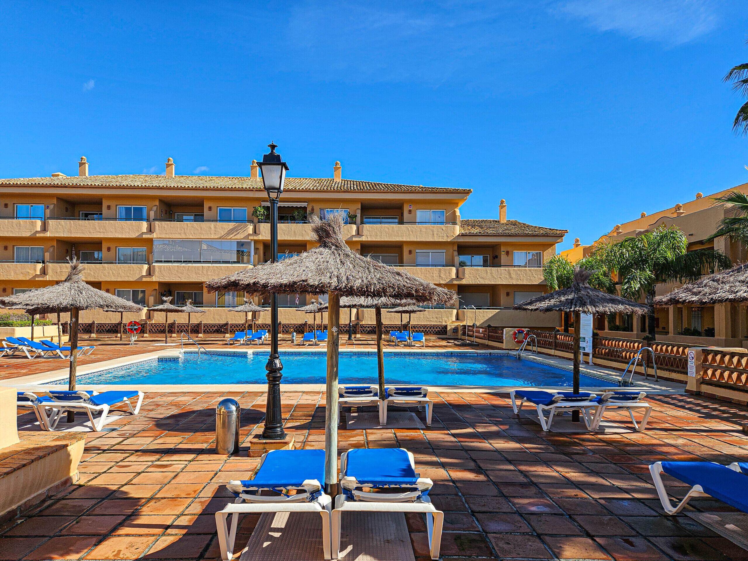 Zona piscina con lettini e ombrelloni, cielo azzurro limpido e un edificio sullo sfondo con numerosi balconi.