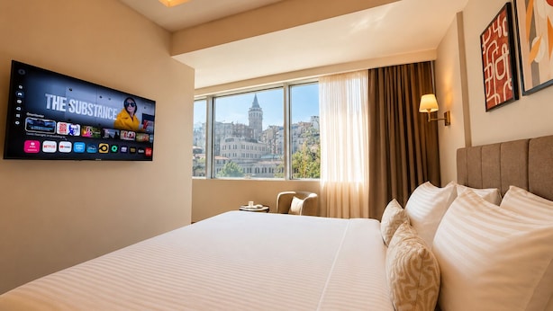 A hotel room with a large bed, a TV mounted on the wall, a window with a city view, and a lamp on the nightstand. - The Tram Karakoy (İstanbul)