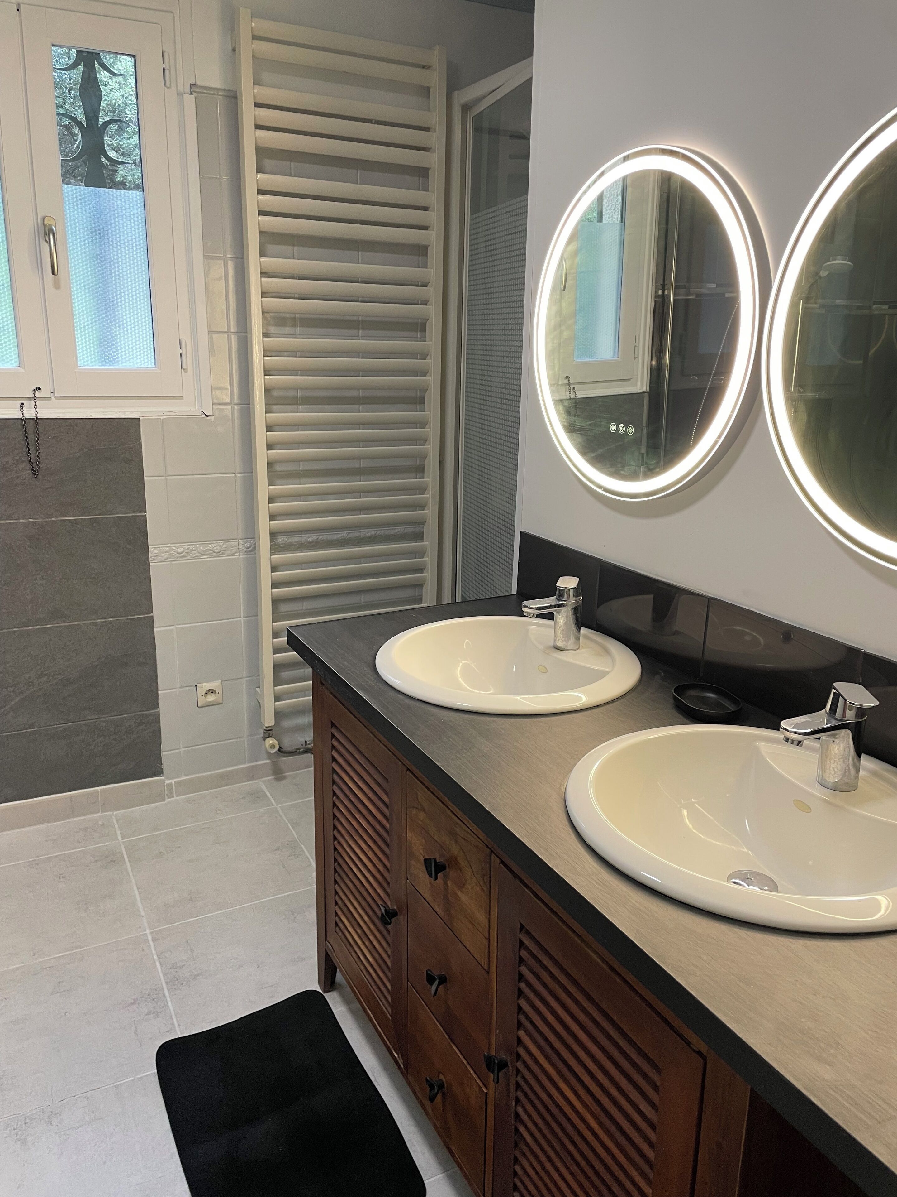 A bathroom with two sinks, a vanity with drawers, a mirror with lights, and a window with blinds.