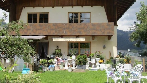 Una casa in legno a due piani con un balcone, immersa in un giardino con piante in vaso e mobili da esterno.
