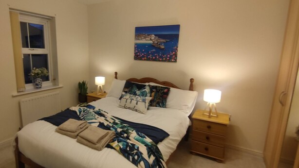 A neatly made bed with a floral-patterned blanket and pillows, a bedside lamp, a small plant, and a window with a view of a coastal scene. - The Parkview House Wakefield - 31 Tuke Grove not 42 Please (Wakefield)