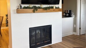 En stue med et fladskærms-tv ophængt over en pejs, et spiseområde med bord og stole, og et køkken med hårde hvidevarer.