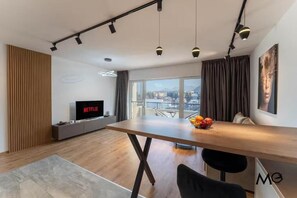 Un salon moderne avec un plancher en bois, une table à manger, un téléviseur et une grande fenêtre donnant sur la ville.