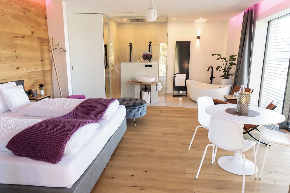 Una habitación de hotel moderna con una cama grande, una zona de estar con una mesa y sillas, una bañera y un ventanal con cortinas.