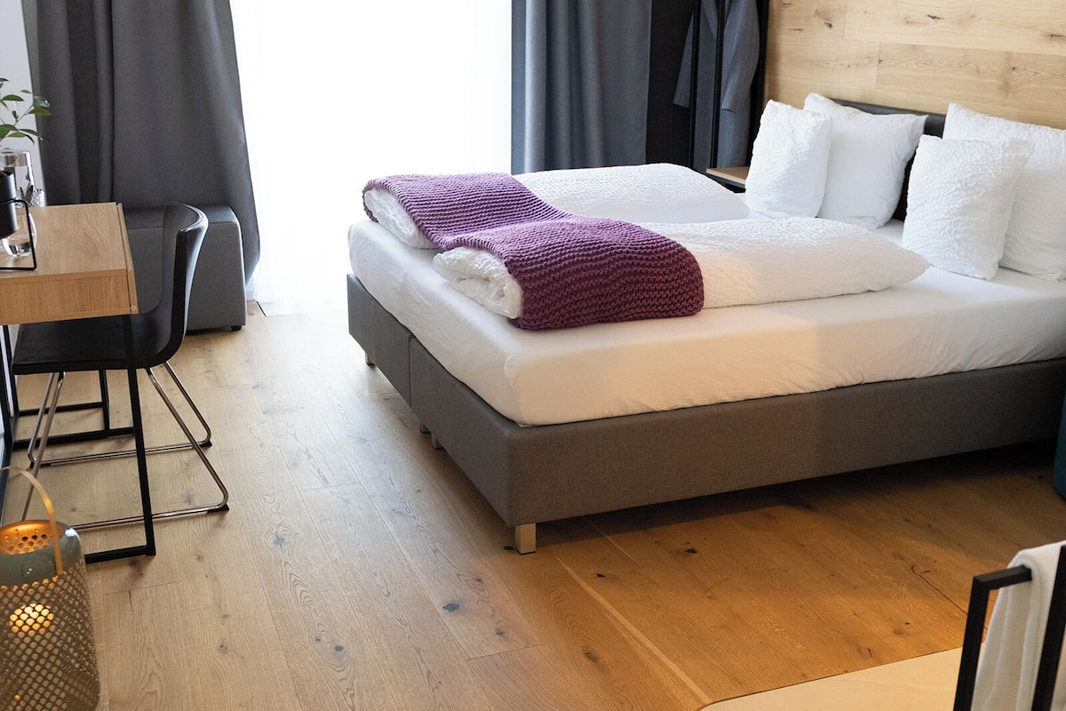 Un dormitorio moderno con cabecera de madera, cama con ropa de cama blanca y morada, escritorio con silla y una ventana grande con cortinas.