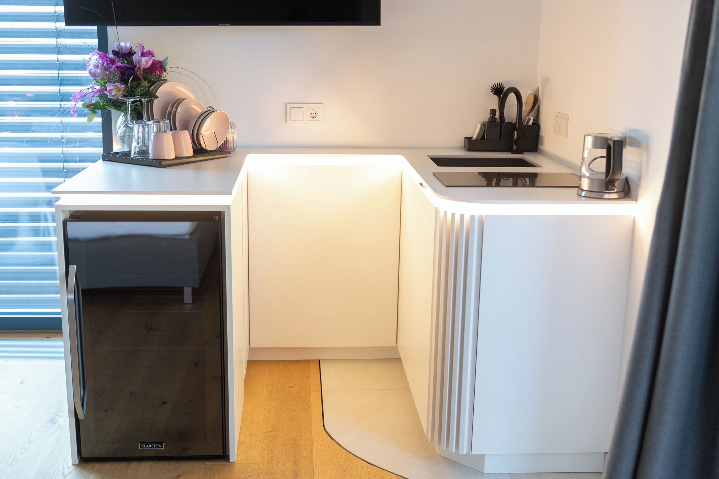 Une cuisine compacte équipée d’un lave-vaisselle, de meubles blancs, d’une machine à café et d’un vase avec des fleurs posé sur le plan de travail.